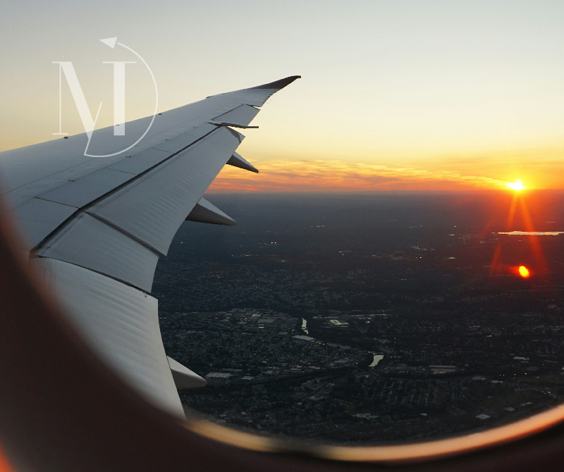 View from a passenger window showing the aircraft wing against a fiery sunset sky above a city, capturing the moment of international arrival and the excitement of air travel.