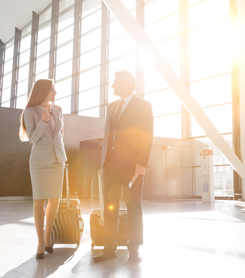 Two business executives in suits, with carry-on luggage, speaking in a modern, sunlit airport terminal, symbolizing efficient corporate travel and seamless global business connections.